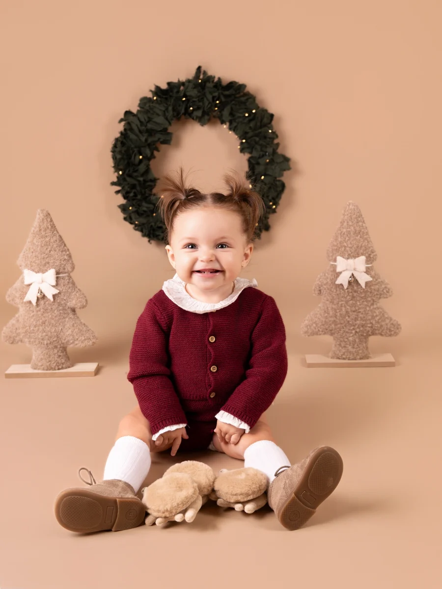 NIña vestida de burdeos sobre fondo marron, con una corona navideña detrás de ella y dos arbolitos. sesiones de navidad 2026