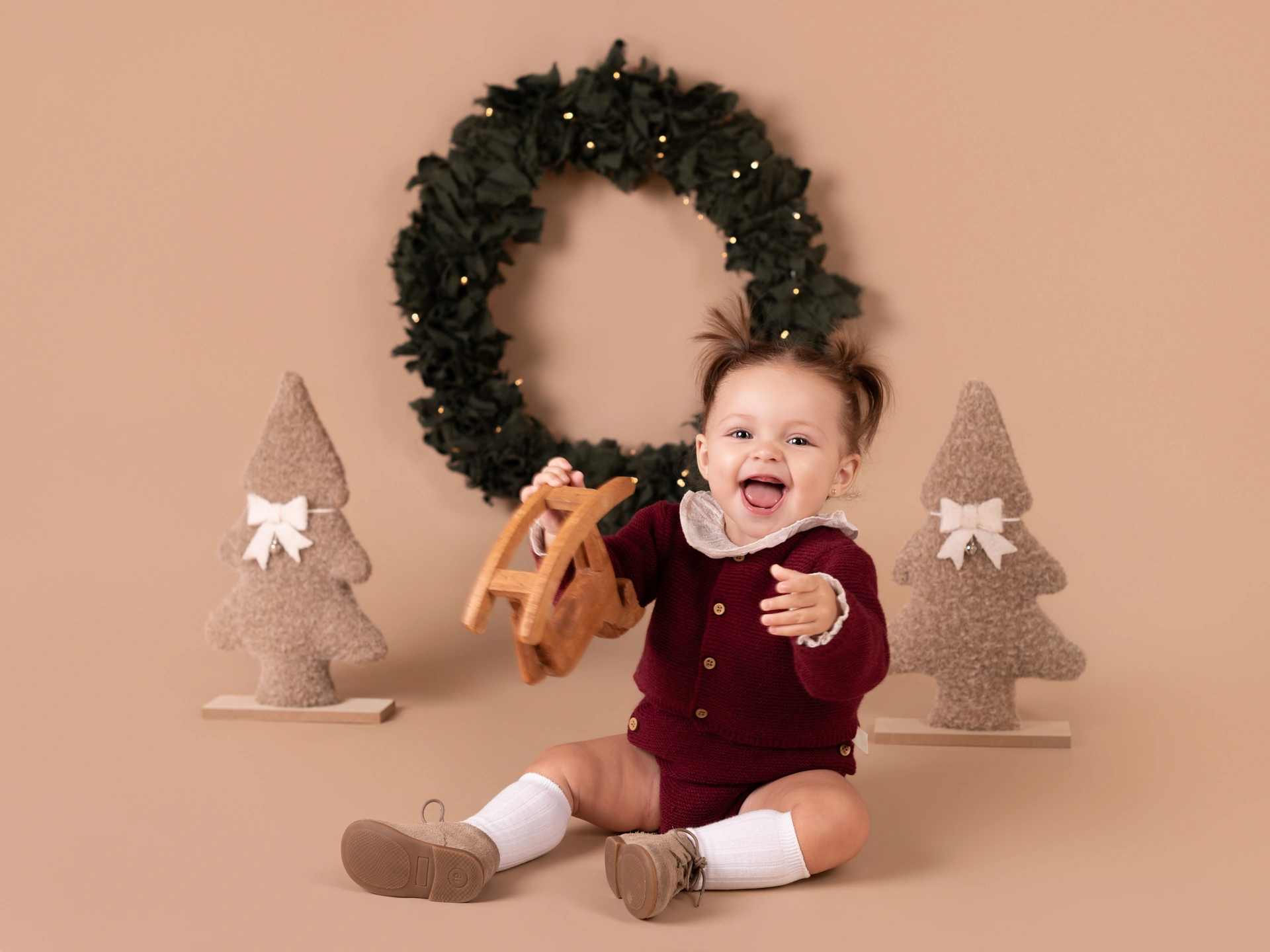 NIña vestida de burdeos sobre fondo marron, con una corona navideña detrás de ella y dos arbolitos. sesiones de navidad 2026